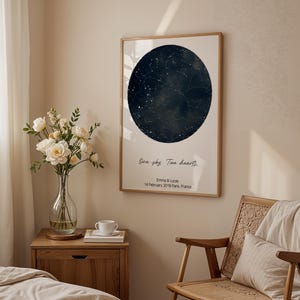 Peut inclure: Impression encadr&eacute;e repr&eacute;sentant une carte c&eacute;leste bleu fonc&eacute; dans un design circulaire. L'impression comprend le texte "One sky. Two hearts." et les noms "Emma & Lucas" avec une date et un lieu. L'impression est affich&eacute;e dans une pi&egrave;ce avec une chaise en bois et un vase de fleurs.