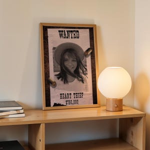 Puede incluir: Un póster enmarcado con el texto "WANTED" y "HEART THIEF" con la foto de una mujer, con una recompensa de 750.000 $. Una lámpara de globo redonda y blanca con base de madera se encuentra en una estantería de madera, junto a una pila de libros.
