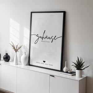May include: A framed black and white print with the word "zuhause" in cursive script, displayed on a white cabinet. The cabinet is adorned with black and white vases, a small plant, and other decorative items. The print includes names.