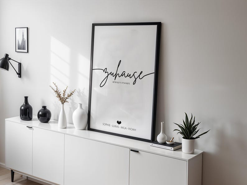 May include: A framed black and white print with the word "zuhause" in cursive script, displayed on a white cabinet. The cabinet is adorned with black and white vases, a small plant, and other decorative items. The print includes names.