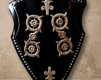 May include: A black shield-shaped wall plaque featuring a border of small white triangular inlays. The center is decorated with two vertical metallic gear-like ornaments and two silver-toned fleur-de-lis symbols positioned at the top and bottom.