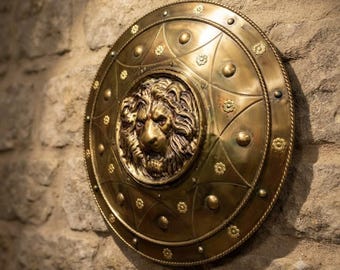 Puede incluir: Un escudo circular de latón que presenta un relieve tridimensional prominente de una cabeza de león en el centro. La superficie está decorada con patrones de estrellas grabados, pequeños tachones circulares elevados y motivos florales.