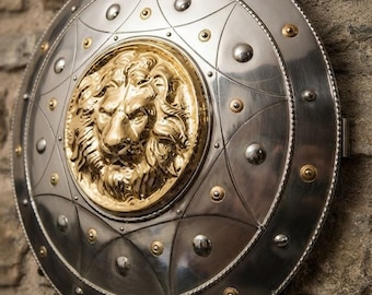 Puede incluir: Un escudo circular de metal con acabado en plata pulida y un relieve de cabeza de león en tono dorado en el centro. La superficie está decorada con líneas curvas grabadas, pequeños remaches plateados y diminutos detalles circulares dorados.