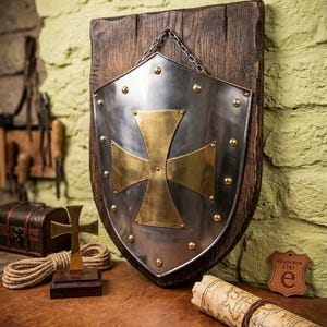 May include: A silver-colored metal shield featuring a large brass-colored cross in the center, secured with brass rivets along the edges. The shield is mounted on a dark, weathered wooden plaque and suspended by a metal chain for wall display.