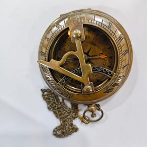 May include: A vintage brass sundial compass with a chain. The compass has Roman numerals around the outer edge and directional markings inside. The gnomon is a curved piece of brass. The compass is on a white surface.