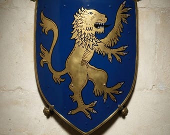 Puede incluir: Un escudo azul con un león rampante dorado, borde metálico y soportes decorativos en las esquinas. Debajo del escudo, una placa rectangular de latón muestra el texto: COAT OF ARMS OF SIR ARTHUR DE BOHUN - C. 1471 HEROIC COLLECTION - EX PRIVATE ESTATE.