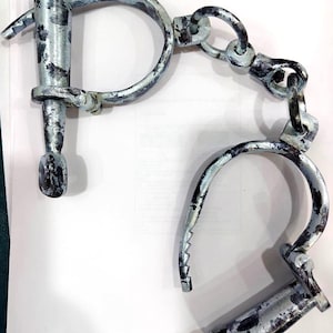 Puede incluir: Un juego de esposas de metal antiguo, que muestra signos de desgaste. Las esposas plateadas tienen pérdida de pintura gris oscuro. El juego incluye dos esposas circulares unidas por una cadena, con un mecanismo de bloqueo y un pasador.