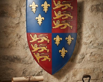 Puede incluir: Un escudo con un blasón cuartelado que muestra flores de lis doradas sobre azul y leones dorados sobre rojo. Una etiqueta debajo dice: Historical Coat of Arms Shield - 14th C. Replica. El escudo está montado en una pared sobre un estante de madera.