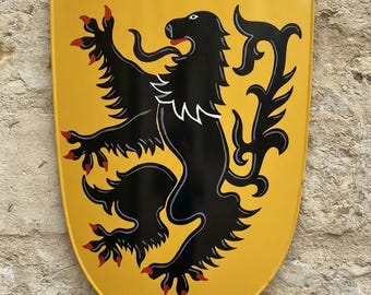 Op de afbeelding: Een geel schild met een zwarte klimmende leeuw met rode klauwen en een rode tong. Het schild heeft een puntige onderkant en een gebogen bovenrand met twee zichtbare metalen klinknagels in de bovenhoeken, voorzien van een heraldisch ontwerp.