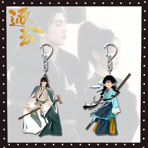 May include: Two anime-style keyrings featuring characters in traditional clothing. One keyring depicts a figure in white and green robes with a staff, the other shows a figure in blue and yellow attire with a staff. Both have silver key rings.