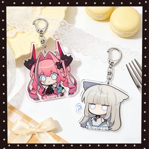 Puede incluir: Dos llaveros de estilo anime con anillas plateadas. Uno presenta un personaje de pelo rosa con detalles negros, la palabra "SAMPLE" está impresa en la parte inferior. El otro tiene un personaje de pelo gris con un signo de interrogación.