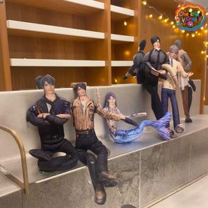 Peut inclure: Des poupées en papier représentant des personnages masculins en tenues variées et une sirène sont disposées sur un banc. Les poupées présentent des vêtements et des traits de visage détaillés, avec des étagères et des guirlandes lumineuses en arrière-plan.