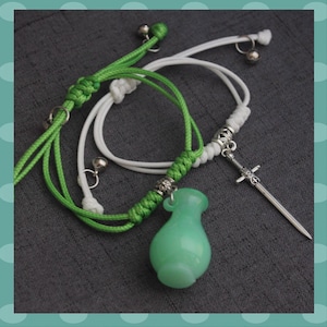 May include: Two bracelets with green and white cords. One has a green vase-shaped charm, the other a silver sword pendant. Both have small silver bells and decorative knots. The background is a grey fabric.