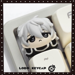 Pode incluir: Uma tecla personalizada com um personagem de desenho animado com cabelo branco e detalhes em preto. A tecla é projetada para caber em um teclado e é cercada por outras teclas brancas. O texto "LOBO KEYCAP" é visível na parte inferior.