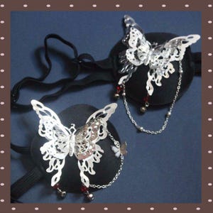 Puede incluir: Dos accesorios para el cabello con forma de mariposa plateada con intrincados diseños calados. Cada mariposa está unida a una base negra y adornada con cadenas colgantes, pequeñas cuentas y detalles rojos. Accesorios para peinado.