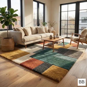 Op de afbeelding: Een kleurrijk vloerkleed met een geometrisch patroon in tinten geel, oranje, groen, grijs en crème. Het kleed ligt in een woonkamer met een beige bank, houten salontafel en een plant in een pot. De kamer heeft grote ramen en een houten vloer.