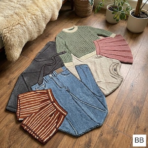 May include: A collection of decorative rugs shaped like clothing items, including a green sweater, a pink skirt, a pair of blue jeans, and a brown and white striped scarf. The rugs are arranged on a wooden floor.