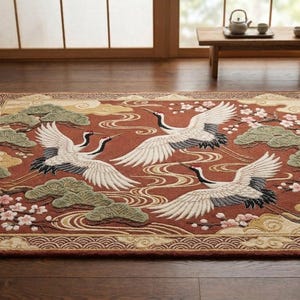 Könnte beinhalten: Rechteckiger Teppich mit japanisch inspiriertem Design. Er zeigt drei weiße Kraniche mit ausgebreiteten Flügeln auf einem rostfarbenen Hintergrund mit stilisierten Wolken, Wasser und floralen Elementen. Der Rand hat ein sich wiederholendes Wellenmuster.