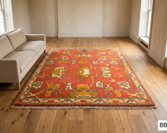 May include: A rectangular, red area rug with an intricate, traditional design. The rug features yellow and white floral and geometric patterns, with a cream-colored border and fringe. The rug is placed on a hardwood floor in a room with a beige sofa.