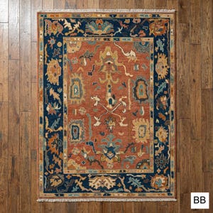 May include: Rectangular area rug with a central terracotta field and a navy blue border. The rug features intricate floral and geometric patterns in shades of teal, gold, and cream. The rug is placed on a wooden floor.