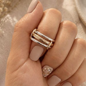 May include: A multi-band ring featuring two silver-toned outer bands and a central gold-toned band, all connected by a small vertical bar on the side. The ring is worn on a finger with matte beige nail polish, with one nail featuring small rhinestones.