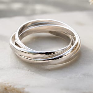 Può includere: Un anello in argento lucido composto da tre fasce intrecciate che creano una forma circolare continua. La superficie metallica è liscia e riflettente, catturando la luce per evidenziare la struttura rotonda di questo gioiello.