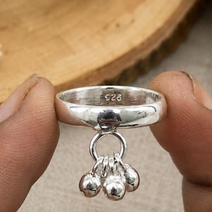 Puede incluir: Un anillo de plata pulida con una banda lisa que presenta el sello 925 grabado en el interior. Un pequeño aro circular está sujeto a la parte inferior de la banda, del cual cuelgan tres diminutos dijes redondos de plata que se mueven.