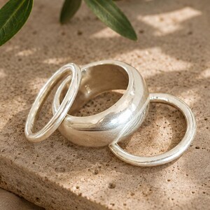 Puede incluir: Un conjunto de tres anillos de tono plateado que consta de una banda ancha y gruesa y dos bandas más delgadas y lisas. Los anillos tienen un acabado metálico pulido y reflectante, dispuestos en grupo para mostrar su forma redondeada.