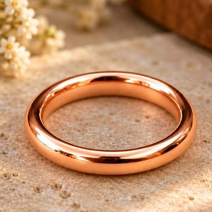 May include: A polished rose gold band ring with a smooth, rounded surface and a high-shine metallic finish. The ring features a simple, continuous circular design with no additional embellishments, stones, or engravings on the metal surface.