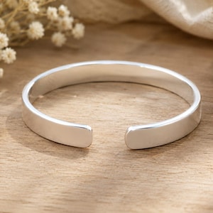 May include: A polished silver cuff bracelet with a smooth, reflective surface and a simple open-ended design. The bracelet features a rounded, minimalist band that curves into a circular shape, showcasing a clean, metallic finish throughout.