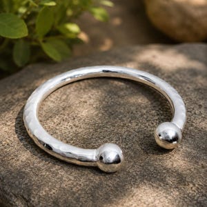 May include: A silver-toned open cuff bracelet featuring a hammered texture along the band and two smooth, polished spherical ball ends. The bracelet has a circular shape with a gap between the two rounded terminals, showcasing a reflective finish.
