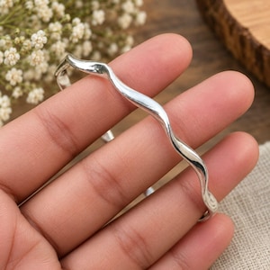 May include: A polished silver-tone cuff bracelet featuring a continuous, smooth, undulating wave design. The metal band has a reflective, metallic finish and a consistent thickness throughout its curved, flowing shape, forming an open-ended loop.