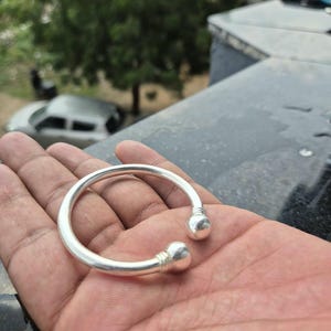 Op de afbeelding: Een zilverkleurige open armband met een gladde, cirkelvormige band die aan beide uiteinden eindigt in gepolijste bolvormige knoppen. Het metalen oppervlak heeft een reflecterende afwerking en bevat subtiele gegroefde details bij de bollen.