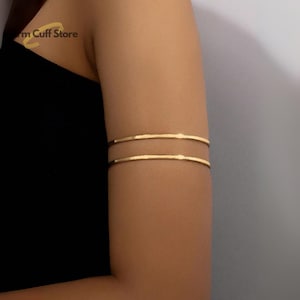 Puede incluir: Joyería de brazalete dorado. El brazalete presenta dos bandas paralelas con una textura martillada. La joya se lleva en la parte superior del brazo. El fondo es de color neutro.