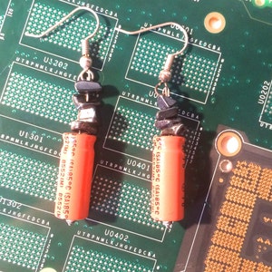 以下が含まれることがあります： オレンジと黒の電子部品で作られたイヤリングのペア。イヤリングは小さなコンデンサと黒いビーズで作られています。イヤリングは銀色のフックにぶら下がっています。