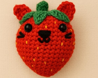 May include: A handmade crochet strawberry cat with a red body, green leaves, and black eyes and nose. The strawberry cat has small ears and yellow seed details. This whimsical item is perfect for a child's room or as a unique gift.