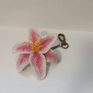 Puede incluir: Un llavero de flor de lirio tejido a ganchillo. La flor tiene pétalos rosas con bordes blancos y estambres amarillos. Está unido a un cierre y cadena de color bronce, perfecto para llaves o bolsos.