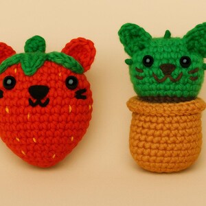 Puede incluir: Dos peluches hechos a mano con ganchillo: una fresa roja con hojas verdes y un cactus verde en una maceta marrón. Ambos tienen ojos negros y caras sonrientes. La fresa tiene pequeñas puntadas amarillas.