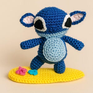 May include: A handmade crochet figure of a blue alien-like character surfing on a yellow surfboard. The character has large ears, black eyes, and a light blue belly. The surfboard has pink and blue flower accents.