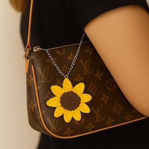 Könnte beinhalten: Eine braune Handtasche mit einem gelb-braunen gehäkelten Sonnenblumen-Anhänger, der an einer silbernen Kette hängt. Die Tasche hat ein sich wiederholendes Logomuster und eine hellbraune Ledereinfassung. Der Hintergrund ist ein schwarzes Oberteil.