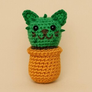 May include: A handmade crochet cactus in a pot. The cactus is green with black eyes and a brown mouth, resembling a cat. The pot is orange. The item is made of yarn.