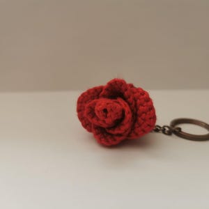 Könnte beinhalten: Ein handgefertigter roter Häkelrosen-Schlüsselanhänger. Die Rose ist detailliert mit sichtbaren Nähten und einem strukturierten Aussehen. Der Schlüsselanhänger hat einen bronzefarbenen Metallring.