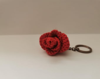 Peut inclure: Un porte-clés fait à la main en forme de rose rouge au crochet. La rose présente des coutures visibles et un aspect texturé. Le porte-clés est attaché à un anneau en métal de couleur bronze.