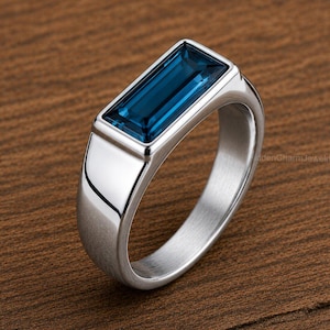 Puede incluir: Un anillo plateado con una gema rectangular de color azul oscuro. El anillo tiene un acabado pulido y un diseño moderno y minimalista. La gema está engastada en un bisel, sobre una superficie de madera.
