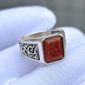 Op de afbeelding: Een zilveren ring met een rechthoekige, roodbruine steen. De ring heeft een sierlijk ontwerp aan de zijkanten. De steen is gezet in een vierkante bezel. De ring is waarschijnlijk gemaakt van sterling zilver.