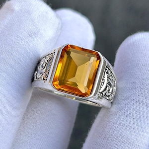Peut inclure: Une bague en argent avec une grosse pierre jaune-orangé rectangulaire. La bague présente des détails complexes sur les côtés. La pierre est la pièce maîtresse, sertie dans un anneau en argent poli. La bague est tenue par une main gantée.