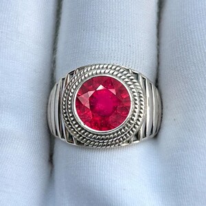 Peut inclure: Une bague en argent ornée d'une grosse pierre rouge vif et ronde en son centre. La pierre précieuse est entourée d'un sertissage texturé et détaillé. La bague a un anneau épais avec des rainures verticales, créant un design audacieux.