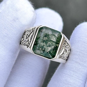May include: A silver ring with a rectangular, moss-green gemstone. The ring has an ornate, engraved design on the band. The gemstone is a vibrant green with natural inclusions, set in a polished silver band.