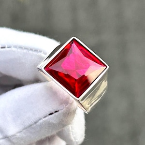 以下が含まれることがあります： シルバーの指輪。大きな正方形にカットされたルビーレッドの宝石が特徴です。宝石は鮮やかな赤色で、磨かれたシルバーのバンドにセットされています。手袋をはめた手で指輪が持ち上げられています。