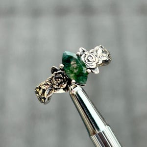 Può includere: Un anello d'argento con una gemma verde taglio marquise. L'anello presenta dettagli floreali sulla fascia, tra cui accenti a forma di rosa. L'anello è tenuto da uno strumento d'argento su uno sfondo sfocato.